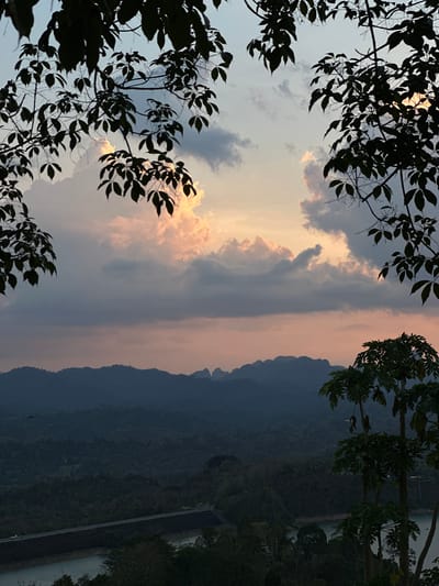 The image captures a landscape in Ban Chiao Lan, Thailand, at what appears to be dusk or dawn. The sky is filled with clouds, some of which are illuminated with a warm, orange light, possibly from the rising or setting sun. Silhouetted tree branches and leaves frame the top and left sides of the image, adding a natural border to the scene. In the background, there is a mountain range. A dam structure with a body of water is visible on the right side. The overall scene conveys a sense of calm and natural beauty.