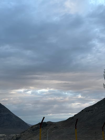 This media file, captured in Chumkar, India, presents a dramatic landscape under a dynamic sky. The scene is dominated by a steep, rugged mountain slope that occupies the right side of the frame, casting a dark shadow. In the foreground, a fence made of barbed wire runs horizontally across the image, with two posts visible – one with red and yellow bands, and another more distant one. The fence suggests a boundary or demarcation in this remote location.
The sky is filled with a complex arrangement of clouds. On the left, the clouds are denser and appear a deep, overcast blue-gray. As the eye moves to the right, the clouds become thinner and more striated, allowing glimpses of sunlight to break through, creating bright streaks and illuminated patches. This suggests either early morning or late afternoon light. The weather appears overcast with pockets of clearer sky, creating a moody and atmospheric effect.
There are no people visible in the image, nor any explicit text. The overall impression is one of natural grandeur and a sense of isolation, emphasized by the imposing mountain and the stark fencing. The interplay of light and shadow on the clouds and the mountain creates a visually striking and somewhat somber mood. The location cues of mountains and the dry, rocky terrain are consistent with high-altitude regions.