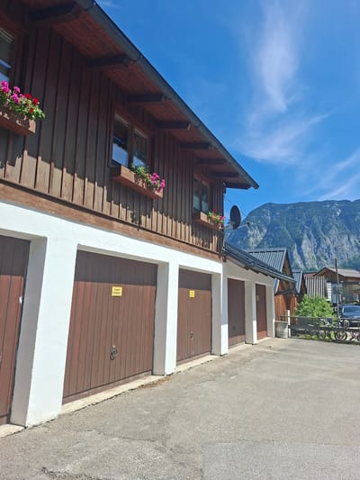 The image shows a row of garage doors on a sunny day in Obertraun, Austria, with a mountainous backdrop. The garages have wooden doors and white trim. Some of the garages have flower boxes with pink and red flowers above them. A satellite dish is mounted on the side of the building. In the background, a steep, forested mountain rises under a blue sky with scattered clouds. To the right of the garages, there is a fence and several bicycles parked. A dark-colored car is also visible. The ground in front of the garages is paved with asphalt. A yellow sign with German text is affixed to one of the garage doors.