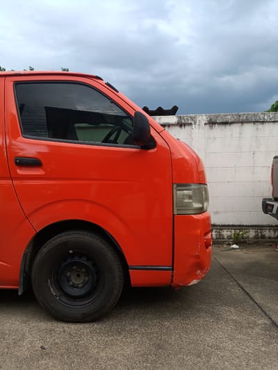 A close-up, side-view of a bright orange van parked on a concrete surface in Ban Patan, Thailand. The van is the primary subject, with its front passenger side and wheel prominently featured. The sky above is overcast with heavy, dark clouds, suggesting an impending storm or a humid, tropical day.
The van shows signs of wear and tear, with a noticeable dent and scuff marks on its front bumper and fender. The windows are tinted dark. A black side mirror is visible, and the door handle is also black. The wheel is a standard black steel rim with a worn tire.
Behind the van, a weathered white concrete wall stretches across the frame. The wall has visible grime and discoloration, indicating age and exposure to the elements. Some debris, including plastic trash, is scattered on the ground near the base of the wall. To the right of the orange van, the rear of another vehicle, possibly a light-colored pickup truck, is partially visible.
The scene appears to be an outdoor parking area or a roadside. The lighting is dim due to the cloudy sky, but the vibrant orange of the van stands out. There is no visible text in the image. The overall mood is somewhat mundane, perhaps even a bit bleak due to the weather and the van's condition, but the bright color of the van provides a focal point. The location cues, such as the wall construction and vegetation, are consistent with a Southeast Asian setting. The time of day is difficult to determine precisely but seems to be daytime.