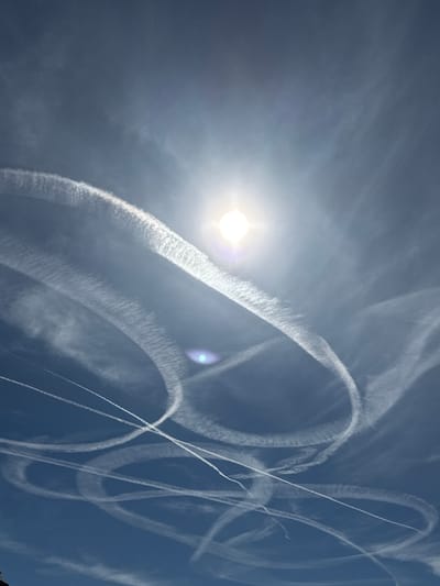 An upward-facing photograph of a bright sun partially obscured by thin clouds and aircraft contrails in the sky above Salach, Germany. The sun is positioned slightly above and to the right of the center of the frame, emitting a strong glare. Numerous white contrails are visible, forming intricate, overlapping arcs and lines across the deep blue sky. Some contrails appear wispy and diffused, while others are more distinct. The overall impression is a busy sky with multiple aircraft having recently passed through.