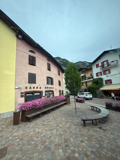 The image shows a view of a cobblestone plaza in Fai della Paganella, Italy. On the left, a pink building has the sign "CASSA RURALE" above its entrance. A long planter box filled with pink flowers runs along the front of the building, with a wooden bench in front of it. To the right of the pink building, a white SUV is parked. Further to the right, a building with a white facade and red shutters has a sign that reads "Bar Locanda Ca'". A person in a white shirt and black pants is walking near an awning with yellow and white stripes. In the background, mountains are visible under a cloudy sky.