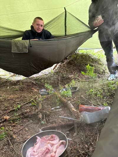 The image shows a camping scene in Macclesfield Forest, United Kingdom, at the coordinates 53.26 latitude and -1.991 longitude. A young man is lying in a hammock under a green tarp. He is wearing a dark blue jacket with a light blue zipper and a black shirt underneath. He is looking directly at the camera with a smile on his face. Beside the hammock are various camping items including a black frying pan with bacon cooking in it, a silver pan, a black bowl, and a red bottle. A second man is partially visible in the image. He is standing and wearing a dark grey hooded sweatshirt. He is holding a white cell phone in his right hand and looking toward the left of the image. The ground is covered with brown branches and green foliage. The environment is a dense woodland area. The scene is lit by natural light, and the overall tone is bright and casual. The image captures a typical camping scene, with the focus on the man in the hammock and the cooking bacon.
