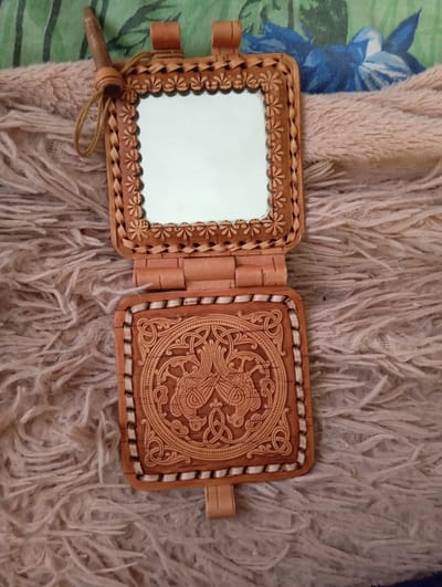 This media file shows a close-up, overhead shot of an open, handmade leather compact mirror. The mirror is in the upper portion of the image, framed by an intricately carved leather border that features a braided pattern and a decorative floral motif along the edge. A thin, light brown cord is attached to the top left of the mirror frame, with a small wooden toggle at its end. The mirror itself is square and reflects a white light, indicating it is clean and functional.
The bottom portion of the compact features a larger, ornately carved leather panel. This panel is square and displays a detailed, intricate Celtic knotwork design with animal figures, possibly depicting birds or dragons. The carving is deep and textured, with a light brown leather background. The edges of this panel are trimmed with a contrasting white stitching and a decorative leather inlay. The bottom of the lower panel has a small leather tab, likely for securing the compact when closed.
The compact mirror is resting on a soft, fluffy, light brown blanket or rug, which occupies the majority of the background. To the left of the mirror, a strip of green fabric with dark stripes is visible. In the upper right corner, a hint of a blue and white floral pattern on fabric can be seen. The lighting suggests it is daytime, possibly indoors, given the even illumination and absence of harsh shadows. No people are visible in the image. The object appears to be an antique or artisanal item due to its detailed craftsmanship. There is no visible text in the image.