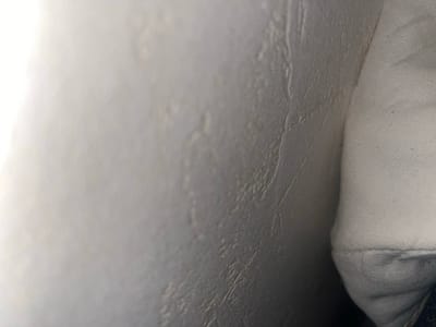 This image presents an extreme close-up of two juxtaposed textured surfaces, located within Clermont-Ferrand, France. The left section of the frame features a light grey, semi-rigid surface exhibiting irregular undulations, fine linear creases, and scattered raised bumps. This surface appears illuminated from the left, creating a subtle gradient of light and shadow. The right section of the frame displays an off-white textile, characterized by a distinct diagonal weave pattern, typical of canvas or coarse cotton. Minute, dark fibrous particles are sparsely visible on the fabric. A prominent horizontal fold or seam runs along the lower edge of this textile. No human figures, identifiable objects beyond the textures, or specific actions are depicted.