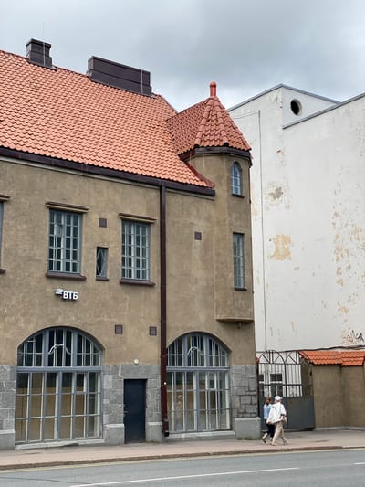 In the city of Sortavala, Russia, a brown building with a terracotta-tiled roof stands to the left, featuring large arched windows on the ground floor and rectangular windows on the upper levels. A prominent sign displaying "ВТБ" is visible on the facade. To the right, a tall, stark white building with peeling paint rises, contrasting with the older architecture. The two buildings are separated by a narrow gap. A black metal gate with decorative elements is present between the buildings, leading to an unseen courtyard. Two women are walking on the sidewalk in front of the buildings, their backs partially to the camera, one wearing a white cap and the other a blue jacket. The sky above is overcast and grey.
