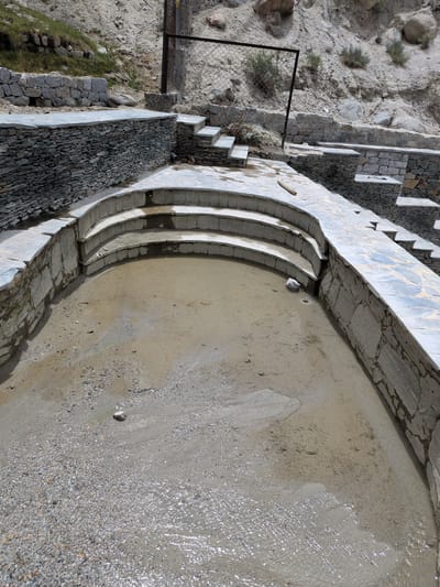 The image shows an outdoor scene in Panamik, India, featuring a partially filled, tiered pool made of stone and concrete. The pool has a curved shape with several descending steps. The water in the pool is shallow and murky, with visible sediment and small pebbles. The surrounding area is constructed with stone walls and flagstone paving. In the background, a rocky, arid hillside is visible, with sparse vegetation. A metal fence structure is also present in the background, bordering the hillside. The overall impression is of a rustic, unfinished, or natural water feature in a mountainous environment.