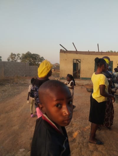 A group of people, including children, stand outside a building with a partially damaged roof. A young boy in a dark jacket occupies the foreground, looking directly at the camera with a serious expression. Behind him are other individuals, one wearing a yellow head covering and another in a yellow top and black skirt. The setting appears to be a rural area with a wall and some trees in the distance. The overall light suggests either dawn or dusk.