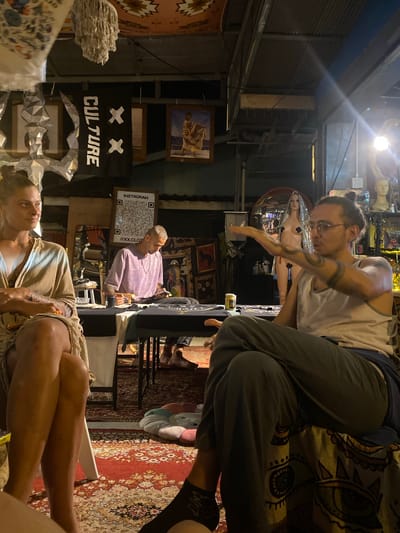 The image depicts an indoor scene with several people and objects, likely in Baan Nai Suan, Thailand.
Foreground: A person lies on a chair or surface, seemingly relaxed. Nearby, another individual sits on a higher surface.
Midground: A person with short, light hair is seated, possibly attending to a task. A banner or sign displaying "XX CULTURE" is visible. A QR code and the word "INSTAGRAM" are displayed on a rectangular sign, with what appears to be the social media handle "@XX.CLOTHING" listed below.
Background: Numerous framed photos or artworks are visible on the walls. The space appears cluttered with various decorative items.
The overall lighting is moderate, casting shadows in certain areas. The setting suggests a shop or exhibition space in Baan Nai Suan, Thailand. The environment hints at artistic or cultural activities.
