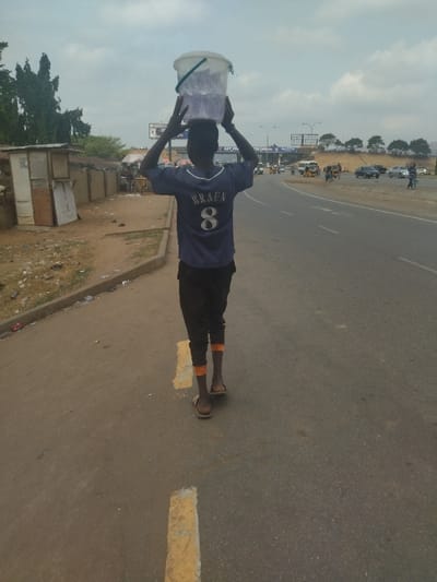 A young man walks along a paved road in Maraba, Nigeria, carrying a white bucket on his head. He wears a blue and white jersey with the word "BRAZA" and the number "8" printed on it. He wears black pants and orange socks, and has a light brown complexion. He is walking in the direction of the city center, towards a busy intersection. There are billboards and signs visible in the distance, and several vehicles can be seen in the background, along with some pedestrians on the opposite side of the road. The street is asphalt with a yellow dividing line.  There are several small buildings on the right side of the image, but they are mostly obscured by trees. The scene is a typical urban landscape in a developing country.