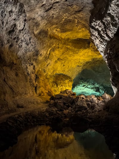 The image presents a dramatic upward view from within a large, illuminated cave or grotto, revealing its rugged rock formations. The scene is characterized by striking artificial lighting that bathes different sections of the cave in vibrant colors. A prominent area on the left and central-upper part of the frame glows with a rich golden-yellow light, emphasizing the intricate textures and vertical contours of the rock walls. Deeper into the cave, towards the upper right, another section is illuminated by an ethereal turquoise-green light, hinting at further passages.

On the far right, a calm body of water, likely an underground lake or pool, reflects the golden-yellow light from the opposite wall, creating elongated, shimmering vertical patterns that resemble dripping stalactites or a watery curtain. The ceiling and portions of the lower left remain in natural, darker rock tones, providing a stark contrast to the brightly lit areas. The raw, uneven surfaces of the rock are visible throughout, showcasing the natural geology.

No people or explicit activities are depicted, but the deliberate lighting suggests this is a managed site for visitors, such as a show cave. There is no visible text. The image evokes a sense of wonder and awe, capturing the mysterious beauty of a subterranean environment, consistent with cave systems found in areas like Haría, Spain.