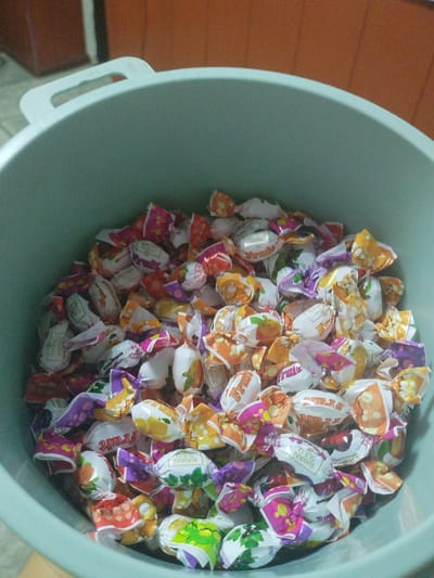 A light blue plastic tub is filled with numerous individually wrapped candies. The candies are colorful and feature various fruit designs, including oranges, lemons, cherries, and grapes, along with the text "Fruit" and "Bayan Sulu". The tub itself has two handles on opposite sides. The background consists of a blurred wooden surface and a white vertical element. The scene appears to be an indoor setting in Mady, Kyrgyzstan.