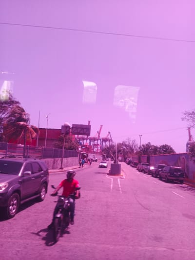 The image captures a street scene in Maiquetía, Venezuela, heavily tinted with a pinkish-purple hue, possibly due to lighting conditions or image processing. A man in a red shirt rides a motorcycle in the foreground, casting a long shadow. To his left, a dark-colored SUV is parked. The road stretches into the distance, lined with several parked cars and a few pedestrians walking on the sidewalk. In the background, large industrial cranes, typical of a port area, are visible, suggesting the proximity to a shipping terminal. A palm tree with wind-swept fronds adds a tropical element to the scene. The sky is a light purple, with some reflections on what appears to be a glass surface, possibly from a vehicle window. The overall impression is of a bright, sunny day, but the unusual color cast gives the image an ethereal quality. There are no discernible signs with visible text.