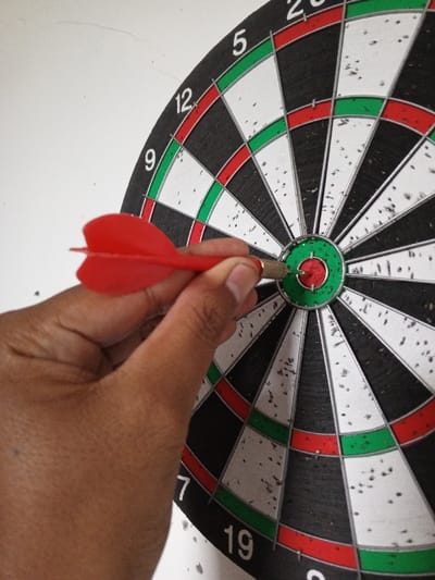 This close-up image, captured in Al Haouafate, Morocco, depicts a human hand manipulating a dart embedded in a dartboard. The hand, exhibiting a medium-dark skin tone, grips a red-finned dart by its metallic barrel and plastic flights. The dart's sharp point is precisely lodged within the red inner bullseye of the circular dartboard. The dartboard itself features concentric rings and radial segments colored black, white, red, and green, with numerical scores visible, including '5', '12', '9', '7', and '19'. The board's surface displays numerous small perforations and impact marks, particularly on the white segments, indicative of extensive use. The background consists of a plain white wall with minor surface imperfections and a faint hairline crack visible in the upper left portion of the frame.