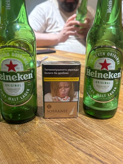 Two green glass bottles of Heineken Original Pure Malt Lager are prominently displayed on a wooden table. One bottle is on the left and the other on the right, both facing forward. Between the bottles sits a pack of Sobranie cigarettes, which has a graphic image of a woman on it and text in a Cyrillic alphabet. The Heineken bottles have distinctive red star logos and their respective labels are visible. In the blurred background, a person wearing a white t-shirt is partially visible, holding another beer bottle. The scene appears to be indoors, with the wooden table suggesting a casual setting. The image does not provide any information about the specific location in Boston, United Kingdom.