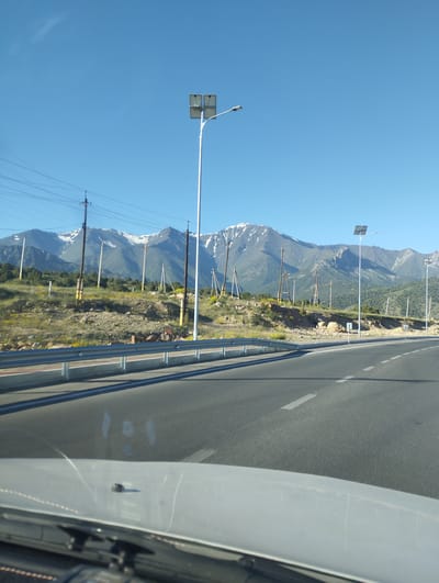 The image captures a scenic view of a mountainous landscape under a clear blue sky, likely taken from a moving vehicle. The foreground shows the hood and windshield of a car, with the road stretching out ahead. A guardrail lines the side of the road. On the slope of the mountain, there are numerous utility poles and some solar-powered streetlights. The mountains themselves are majestic, with snow-capped peaks visible in the distance, and the slopes are covered in green vegetation and sparse yellow wildflowers. Power lines are strung between the poles, crisscrossing the scene. The overall impression is one of a rural or semi-rural setting, possibly on the outskirts of a city or town. There are no people visible in the image. Based on the visual cues, the location could be in or around Myk, Uzbekistan, given the typical mountainous terrain and infrastructure seen in such regions.