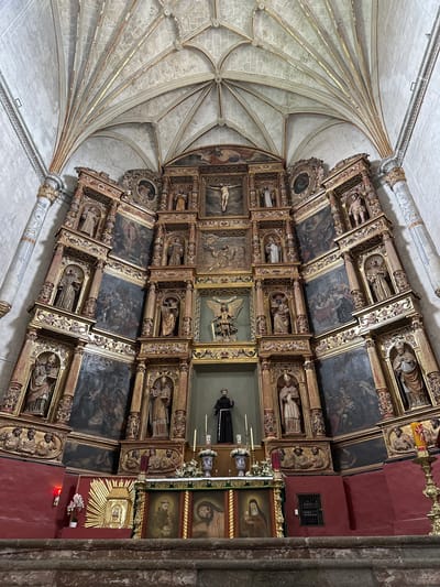 The image shows the interior of a church in Huejotzingo, Mexico, specifically focusing on a large, ornate altarpiece. The altarpiece is highly decorated with gilded wood, featuring numerous niches holding statues of saints and religious figures. Painted panels depicting biblical scenes are integrated into the structure. In the center of the altarpiece, a figure of Christ on the cross is prominently displayed. Below this, a statue of a robed male figure stands in a niche. The ceiling of the church is visible at the top, displaying intricate ribbed vaulting with gilded detailing. In the foreground, the altar itself is visible, adorned with paintings and religious artifacts, including candles and floral arrangements. The walls of the church are painted a deep red. The overall scene conveys a sense of historical and religious significance.