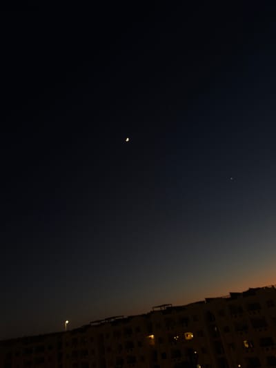 The image shows a nighttime view of a city skyline at dusk, likely in Sidi Abdel Rahman, Egypt, given the context. The sky is a deep gradient of dark blue to a lighter, warm hue near the horizon, suggesting the last vestiges of daylight. A crescent moon is visible in the upper portion of the sky. To the right of the moon, a smaller, brighter point of light, possibly a planet or a star, is also present. Below the sky, a silhouetted building stretches across the lower half of the frame. The building appears to be a multi-story structure with visible windows, some of which are illuminated with warm light. A single streetlamp is illuminated on top of the building, casting a bright light upwards. The overall scene depicts an urban environment at twilight, with celestial bodies above and the architectural features of a city below.