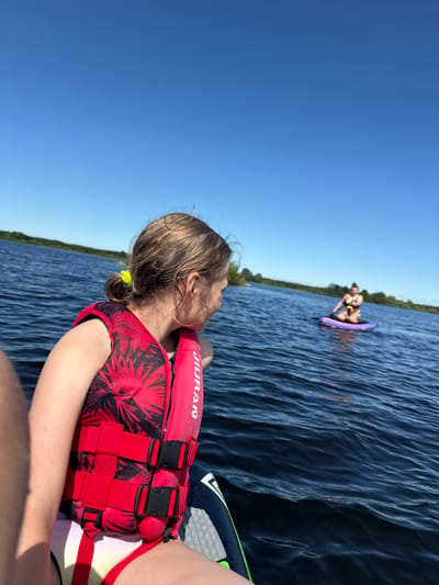 A young girl in a red life jacket with a palm leaf pattern is sitting on a wakeboard or similar watercraft, looking to her right. She has blonde hair tied back with a green scrunchie. Another person is visible in the distance on a purple float in the blue water. The scene is a lake with a tree-lined shore in the background, under a clear blue sky. The sun appears to be high in the sky, suggesting it is daytime. The water is calm with ripples. The text "JIURAN" is visible on the girl's life jacket. The overall mood is recreational and summery.