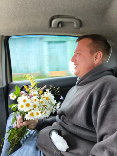 A man with short, light brown hair is sitting in the back seat of a car, holding a bouquet of daisies and wildflowers. He is wearing a grey hooded sweatshirt and blue jeans. The man is smiling and looking to his right, out the car window, where a blurry blue and green landscape is visible. The interior of the car is visible in the foreground and background, with a grey headliner and a dark grey car door. The image appears to have been taken in Medvedyovka, Russia, based on the surrounding landscape.