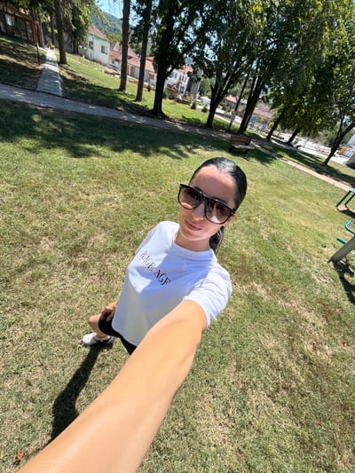 In the city of Chavdartsi, Bulgaria, on a sunny day, a woman stands in a grassy park. She is wearing a white t-shirt with "RAGE AGF" printed on it, black shorts, and white sneakers. Large, dark sunglasses obscure her eyes, and her dark hair is pulled back into a ponytail. She is holding a phone to take a selfie, her arm extending into the foreground. The park features a paved pathway, trees, benches, and children's playground equipment in the background. Residential buildings are visible behind the trees. The grass is somewhat dry, indicating a warm season.