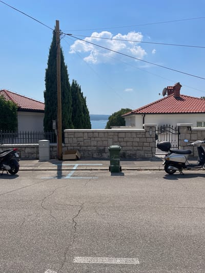 In Crikvenica, Croatia, on a sunny day, a street scene unfolds with a paved road in the foreground showing some cracks. Two scooters are parked on the roadside. To the left, a black scooter is partially visible, while to the right, a silver scooter is parked in front of a white house with a red-tiled roof. A green trash bin stands on the sidewalk next to the house, and a cardboard box is placed on the ground nearby. Behind the sidewalk, a stone wall separates the street from a property with a fence and tall cypress trees. In the distance, beyond the houses, a body of water is visible under a clear blue sky with scattered clouds. A wooden utility pole with overhead wires stands prominently in the middle ground.