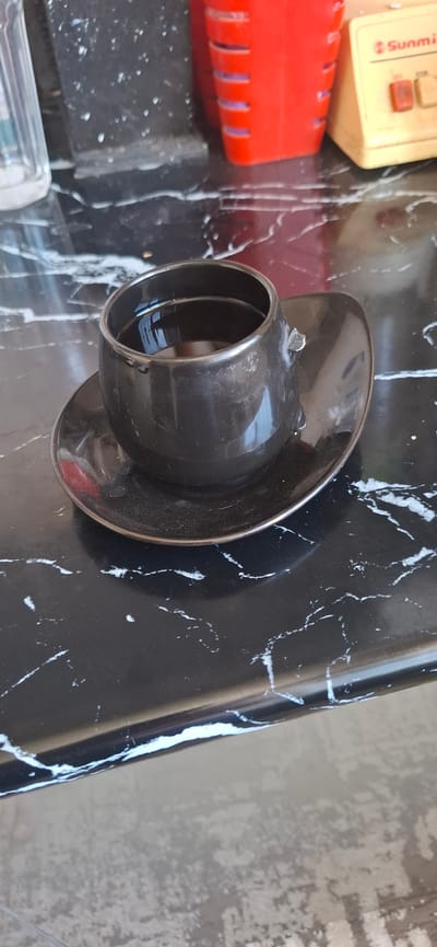 The image shows a close-up of a dark, metallic object with a flared end, resembling a funnel or nozzle. It is positioned on a black, reflective surface with a marbled pattern. To the left of the object, there are hints of other items, including a bright red, ribbed plastic object and a cream-colored rectangular device with some buttons. A small amount of liquid or condensation is visible on the marbled surface, creating streaks and reflections. The lighting suggests it might be indoors with overhead illumination, casting subtle shadows. There is no discernible activity or event taking place; the scene appears to be a still life arrangement of objects. No people are visible. The overall impression is of a detail shot focusing on the texture and form of the metallic object and its surroundings. Visible text on the cream-colored object is partially obscured but includes the word "Sunmi". The location context of Karmana, Uzbekistan, does not provide specific visual cues within the image itself.
