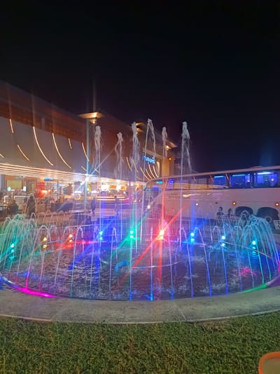 The image depicts a brightly lit fountain at night in Thap Sakae, Thailand. The fountain is round, with water jets illuminated in various colors including red, green, blue, and purple. Behind the fountain is a modern building with a sign in Thai script above its entrance. There is a bus parked to the right of the fountain. Some people are standing near the bus. The sky is dark. The fountain is surrounded by greenery. The lighting on the building casts a warm glow, contrasting with the colorful fountain. There are chairs and tables visible in front of the building suggesting dining or seating area.
