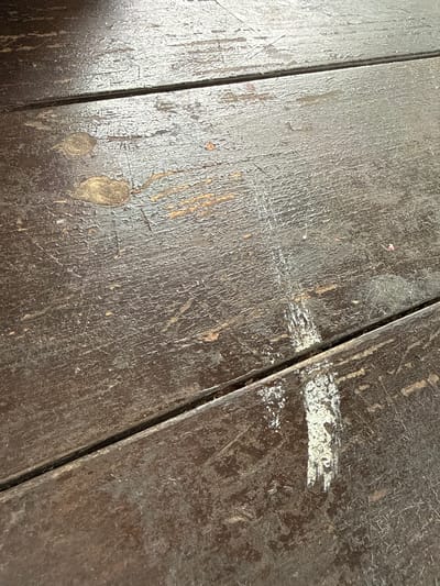 The image shows a close-up, angled view of a dark wooden surface, likely a table or floor, with visible wear and tear. The wood is composed of planks joined together, with a distinct gap between two of the planks running diagonally across the frame. The surface exhibits a rough texture, with peeling paint, scratches, and some lighter-colored patches. There are also circular marks and a vertical streak of white material on the wood. The lighting suggests an indoor environment. The image does not depict any people, specific actions, or interactions. It is a detailed, forensic depiction of a worn wooden surface. The location in Leninskii, Russia, is not discernible from the visual information in the image.