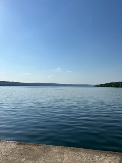 The image shows a wide, calm river with a clear blue sky. The foreground features a concrete embankment. In the distance, a treeline marks the opposite bank, with hints of buildings visible on the right side of the frame. The water has gentle ripples. The sky is mostly clear, with a few wispy clouds. The overall impression is one of a serene natural landscape, possibly within or near the city of Korzhevo, Moldova, given the contextual information.