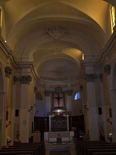 The image shows the interior of a church, likely the Basilica di San Marino, in the City of San Marino, San Marino. The architecture is ornate, with a vaulted ceiling, large pillars, and decorative molding. A crucifix is prominently displayed at the altar, which is adorned with religious artwork and candles. Rows of wooden pews are visible in the foreground, indicating a space for congregants. The lighting is dim, with warm tones suggesting artificial illumination. There are no people visible in the image.