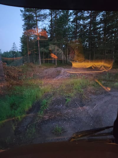 This image, captured from the interior of a vehicle, shows a rural forest scene at dusk. The view is through the windshield, which has some reflections, including an orange and yellow light source and a hint of the vehicle's interior. The foreground consists of a dirt and gravel road with some green grass and vegetation on the left. In the midground, there is a cleared area with piles of felled trees and debris, indicative of logging or forest management. Beyond this, a dense forest of tall pine trees lines the horizon. The sky is a dusky blue. The presence of felled trees and the general setting suggest this location could be a rural area outside of the city center of Lohja, Finland, possibly near a logging site or a managed forest. There are no people visible in the image.