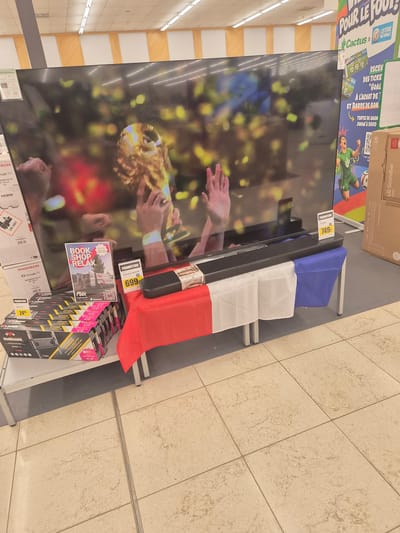 The image shows a retail display featuring a large television screen and various electronic accessories. The television displays a celebratory scene, possibly related to a sporting event, with confetti and hands reaching out. To the right of the television, a soundbar is displayed, draped with the French flag. Below the soundbar and television, there are boxes of electronic accessories, including soundbars and possibly television mounts.
Visible text includes brand names like "LG Soundbar," "THOMSON," "meliconi," and "Cactus." Promotional text such as "POUR LE FOOT!", "PROMOTION," and prices like "749:" and "699:" are also present, indicating a sales event. A booklet titled "BOOK. SHOP. RELAX." is also visible.
The setting appears to be an electronics store, suggested by the display shelving, lighting, and tiled floor. The overall impression is that of a commercial display, likely in a supermarket or electronics retailer, possibly during a promotional period related to a sporting event like the World Cup, given the football imagery and the French flag. The lighting suggests an indoor environment. No specific time of day or weather conditions can be determined.