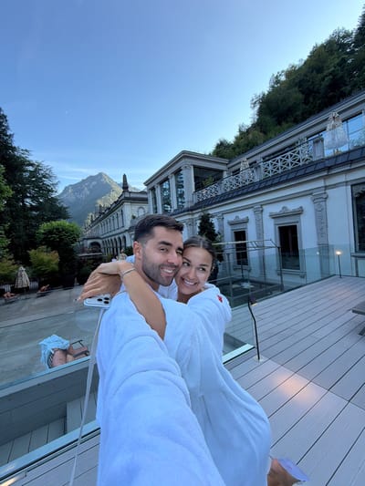 A couple, a man and a woman, are taking a selfie outdoors in San Pellegrino Terme, Italy. They are both wearing white bathrobes. The man is on the left, with his arm around the woman. The woman is on the right, with her arm around the man. They are both smiling. In the background, there is a large, ornate building with columns and balconies. There are also trees and a mountain in the distance. The sky is blue. The couple is standing on a wooden deck with a glass railing. There are lounge chairs and umbrellas visible in the background. A phone is visible in the man's hand, used to take the selfie.
