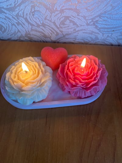 A close-up, eye-level shot shows two rose-shaped candles and a heart-shaped candle on a pink and white oval tray. The rose candles are lit, with flames flickering. The left rose is a creamy yellow color, while the right rose is a bright pink. The heart-shaped candle is a textured red. The tray sits on a wooden surface, and a textured wallpaper with a floral pattern is visible in the background. The image was taken in Kurskaya, Russia.
