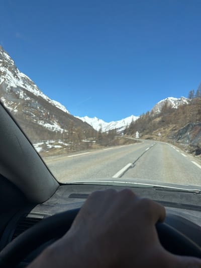 The image is taken from inside a vehicle, likely a car, driving on a paved road. The road winds alongside a mountain range. The mountains are partially covered with snow. The sky is clear and blue. A person's hand and a portion of the steering wheel are visible in the foreground, suggesting that the person is driving. Based on the landscape and context clues, this image was likely taken near Le Monêtier-les-Bains, France.