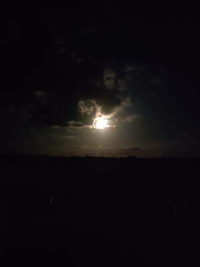 A nighttime photograph captures a partially obscured moon or sun, casting a bright glow through dark, swirling clouds. The lower portion of the image is dominated by a silhouette of a dark landscape, likely fields or trees, creating a stark contrast with the illuminated sky. The scene appears to be an outdoor setting in Cherevky, Ukraine, under a dramatic nocturnal sky.