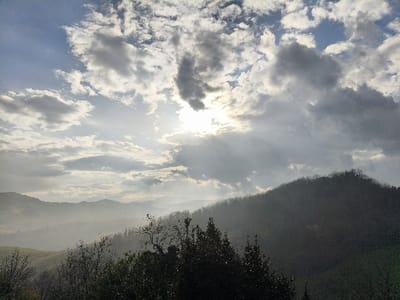 This image depicts a mountainous landscape in Villa di Gemmano, Italy, under a partially overcast sky. The sun, positioned in the upper left quadrant, is partially obscured by bright, diffuse clouds, creating a strong backlight and rays of light penetrating through gaps in the cloud cover. The sky exhibits a mix of bright white and grey clouds, with patches of clear blue visible. In the foreground, the tops of several deciduous trees, some with bare branches and others with foliage, are visible, silhouetted against the brighter midground. The midground and background consist of multiple layers of rolling hills or mountains, heavily forested, receding into a prominent atmospheric haze or mist that softens their outlines and creates a sense of depth. The terrain appears rugged and vegetated. No people, man-made structures, or overt actions are discernible in the scene. The overall lighting suggests either early morning or late afternoon.