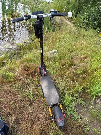 The image shows an electric scooter parked on the grassy bank of a body of water, likely a lake or pond. The scooter is black and silver with red accents on the suspension. It has a display screen on the handlebars and a small black bag attached. In the background, tall reeds grow along the water's edge, and lush green bushes and trees form a backdrop. The ground is covered in a mix of green and dry grass. The scene appears to be in a natural setting, possibly a park or recreational area. The location is identified as Kirzinė, Lithuania. There are no people visible in the image.