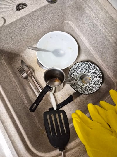 An overhead view within a kitchen sink in the Municipal Unit of Corinth, Greece, reveals a collection of used dishware and utensils awaiting cleaning. The sink is a speckled, light gray composite material with a circular metal drain visible in the upper left corner. Positioned in the sink are a white ceramic bowl containing a silver spoon, a black and white patterned ceramic plate with a silver fork resting on it, a small silver metal saucepan with a black handle, an additional silver knife and fork, a black slotted plastic spatula, and a small white object resembling a scrubber or utensil handle. A pair of bright yellow rubber dishwashing gloves are partially visible in the lower right, lying near the accumulated items. The items display evidence of food residue and use.