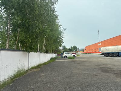 The image depicts an outdoor scene, likely on an overcast day, featuring a gravel parking lot in the foreground. To the left, a white concrete wall runs parallel to the lot, bordered by green grass and some tall reeds. Behind the wall, a dense line of birch trees with white trunks and lush green foliage forms a natural barrier. Further in the background, to the right, is a large, bright orange industrial building with numerous windows. Adjacent to the building, a white tanker truck with multiple axles is visible, partially obscuring a chain-link fence. A white SUV is parked in the gravel lot near the wall, and another darker colored vehicle is behind it. A road with a red and white striped pole is visible in the middle distance. The overall environment suggests an industrial or commercial area on the outskirts of Заслаўе, Belarus. There are no people visible in the image.