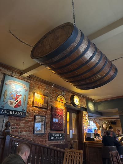 The image shows the interior of a pub in the Vale of White Horse, United Kingdom. A large, wooden barrel hangs from the ceiling by chains. On the brick wall are various signs and framed pictures, including: "MR WARRICKS ARMS", "MORLAND," "YKE BRADY Brewers Cooperage," "HOTEL & RESTAURANT" and one listing a beer house. A clock hangs on the wall. In the background is a view of the bar area with a group of people. A person is visible on the bottom right, standing next to a wooden staircase.