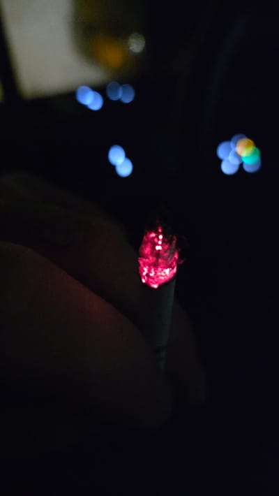 The image displays a lit cigarette in a dark environment. The cigarette's tip is incandescent, emitting a bright red glow from the burning tobacco. It is held by an individual whose fingers are partially discernible in the lower-left portion of the frame. The cigarette paper appears light in color. The background is characterized by significant bokeh, revealing numerous out-of-focus, circular light sources. These lights are predominantly blue, with some yellow, orange, and green points interspersed, indicative of distant artificial illumination, possibly from a city or road at night. The location of this scene is Beringen, Belgium.
