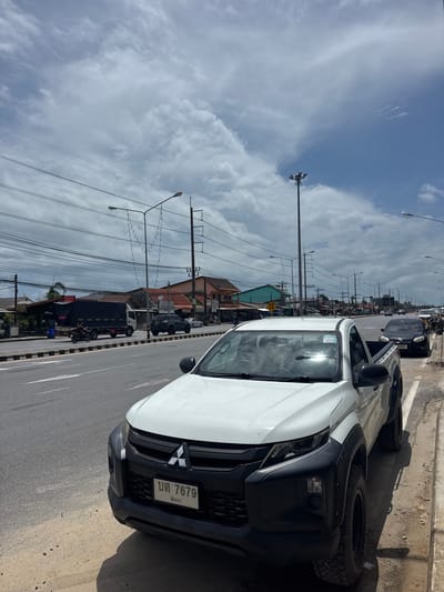 A white Mitsubishi Triton pickup truck is parked on the side of a road in Ban Bang Niang, Thailand. The truck has a black grille and fender flares, and a license plate that reads "บต 7679 พังงา". The road is a multi-lane asphalt road with a sidewalk made of sand and dirt. Overhead power lines and streetlights are visible, as are several buildings along the roadside. In the background, a cloudy sky dominates the scene. Other vehicles, including a black SUV, a white sedan, and a truck with a black tarp, are also present on the road. A person is riding a motorcycle in the distance. The environment appears to be a commercial or mixed-use area in a tropical climate.