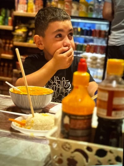 A young boy with dark hair and brown eyes is the primary subject in this media file. He is wearing a black t-shirt with a white graphic design. His right hand is raised to his mouth, and he appears to be eating or about to eat something. His facial expression suggests curiosity or concentration.

The boy is situated in what appears to be a food establishment, possibly a market or a store. In the background, shelves are lined with various products, some of which are in bottles and jars, displaying a range of colors. Two condiment bottles, one with an orange cap and the other a yellow dispenser, are visible in the mid-ground, suggesting a food service setting.

In the foreground, there are two food items presented in small containers. One is a portion of what looks like corn on the cob or a similar snack, with a wooden skewer inserted into it. This is in a ribbed, metallic-colored bowl. Next to it is another food item, possibly a fried snack or a corn dog, served in a white plastic holder, also with a skewer. The lighting suggests it could be daytime, as there is natural light contributing to the scene, though it's difficult to determine the exact time of day. The setting appears to be indoors, with a generally casual and possibly busy atmosphere given the background. No specific weather conditions are discernible. The location context provided is Artik, Armenia, which might imply the type of food or the general environment.