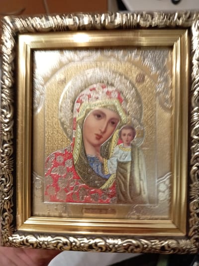 The image shows a framed icon of the Virgin Mary and the Christ Child. The icon is ornate with a golden background and intricate details on the clothing. The frame is also ornate and appears to be gold-colored with embossed patterns. The image is a close-up shot, with the bottom left corner of the frame slightly out of focus. The background of the image is blurred and appears to be an indoor setting with wooden surfaces. The location in the city of Прыгарадны сельскі Савет, Belarus, is not discernible from the image itself.