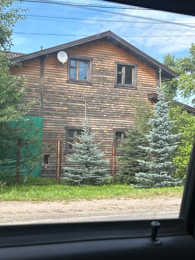 A two-story wooden house, weathered and grey, stands behind a narrow strip of green grass and a dirt road. The house has a pitched roof with dark trim and two windows on the upper level, each divided into four panes. A white satellite dish is mounted on the upper left side of the house. The lower level of the house has two windows with similar panes. In front of the house, three evergreen trees are visible: a medium-sized blue spruce in the center, a smaller dark green evergreen to its left, and a larger, lighter blue spruce on the right. Behind the house, there is dense green foliage and a portion of a rusty metal fence. To the left of the house, a large green tarp is visible, partially obscuring part of the lower wall and fence. The sky above is a bright blue with a few wispy white clouds. Electrical wires are strung across the top of the frame. The image appears to be taken from inside a vehicle, with the edge of a dark interior visible in the foreground. The scene is located in the city of Lotoshino, Russia.