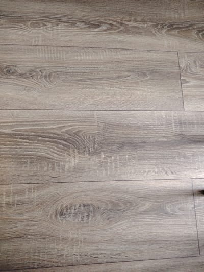 The image displays a close-up, overhead view of a floor with a wood-like laminate finish. The planks are oriented horizontally and are of a uniform width, showing a realistic wood grain pattern with variations in color and texture. There are visible lines between the planks, indicating the seams of the flooring. The lighting suggests an indoor environment, likely in a residential or commercial space. There are no people, animals, or other objects clearly visible in the frame. The texture of the laminate appears to have a matte finish, with subtle details mimicking natural wood knots and grain. The overall impression is of a clean and modern flooring material. Without further context, it is impossible to determine its specific location within Putilkovo, Russia.