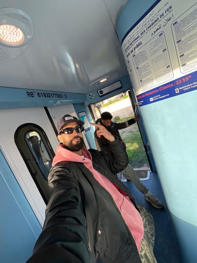 The image shows the interior of a train carriage, viewed from a slightly rotated perspective, as if taken from a selfie-style angle with the camera held by the person in the foreground. The train interior is modern with light blue and white panels.

In the foreground, a man with a beard and sunglasses is visible, looking towards the camera. He is wearing a black jacket over a pink hooded sweatshirt (hoodie) and a baseball cap. His right arm is slightly extended, suggesting he is holding the camera.

Behind him and to his right, another man is visible. He has short, dark hair and is wearing a dark jacket and grey trousers. He is partially obscured, but his right hand is raised, with his index finger pointing forward, and another finger (possibly the middle finger) is also visible. His left hand is also visible, with fingers pointing. He appears to be leaning against the train wall or a seat, looking out a window.

Through the train windows, blurred green foliage and what looks like an outdoor scene with buildings in the distance can be seen, indicating the train is in motion or stopped beside a landscape. The sunlight is bright, suggesting it's daytime.

Above the window line, along the top section of the train wall, there is signage. The text on the signage, partly in Arabic and partly in French, reads:
-   "المكتب الوطني للسكك الحديدية" (National Railway Office)
-   "OFFICE NATIONAL DES CHEMINS DE FER"
-   "Avis au public" (Public Notice)
-   "إعلان للعموم" (Public Announcement)
-   "Centre de Relation Clients: 2255*" (Customer Relations Center: 2255*)
-   "oncf.ma"
-   "oncf-voyages.ma"
-   "Application Mo" (likely "Mobile Application")
-   "ONCF Trafic"
-   A number "RB¹¹ 61932177082-5" is also visible on a lower panel.

The lighting within the train is bright, with some light sources visible on the ceiling panel, including a circular light fixture with multiple small LED-like elements. The overall atmosphere appears casual and relaxed, with the two individuals possibly traveling together.