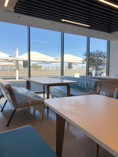 The image captures an unoccupied, modern interior space with panoramic windows overlooking an arid exterior environment, situated in Hanak, Saudi Arabia.
The indoor area features a reflective, light-toned floor and a ceiling composed of parallel black slats alternating with white panels, which integrate recessed spotlights and linear fluorescent fixtures. Large, floor-to-ceiling glass windows, separated by dark vertical mullions, form the main exterior wall.
Furniture within the room includes several light-colored, rectangular tables with rounded corners and wooden legs. Seating comprises individual chairs with wooden frames and dark metal legs, and longer benches upholstered in blue fabric. A significant portion of this furniture, including chairs and benches, remains wrapped in clear protective plastic film, suggesting a new or unutilized status. A segment of an unwrapped blue upholstered seat is visible in the lower-left foreground.
Through the windows, a clear blue sky with a faint contrail is visible. The exterior landscape features a paved area bordered by multiple large, white, tensile-fabric canopy structures, likely serving as sunshades or covered parking. A chain-link fence stretches across the mid-ground, separating the immediate paved zone from more distant, undeveloped terrain exhibiting sparse vegetation. A small tree is visible on the right, partially obscuring a white pickup truck parked in the background. No individuals or discernible activities are present in the scene.